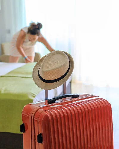 Ankunftsszene in einem Hotelzimmer: Im Vordergrund steht ein orangefarbener Rollkoffer. Auf dem Griff, den man zum Ziehen ausfahren kann, hängt ein heller Männerstrohhut. Im Hintergrund zupft eine Frau mit dunklen, zu einem Knoten zusammengefassten Haaren an der Bettwäsche eines Doppelbettes.