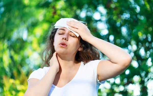 An einem Sommertag steht eine junge Frau unter Bäumen. Die Hitze setzt ihr zu. Sie hat die Augen geschlossen und hält sich ein Tuch an die Stirn.