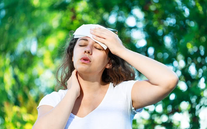 An einem Sommertag steht eine junge Frau unter Bäumen. Die Hitze setzt ihr zu. Sie hat die Augen geschlossen und hält sich ein Tuch an die Stirn.