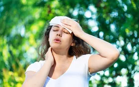 An einem Sommertag steht eine junge Frau unter Bäumen. Die Hitze setzt ihr zu. Sie hat die Augen geschlossen und hält sich ein Tuch an die Stirn.
