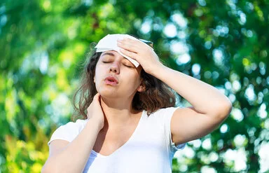 An einem Sommertag steht eine junge Frau unter Bäumen. Die Hitze setzt ihr zu. Sie hat die Augen geschlossen und hält sich ein Tuch an die Stirn.