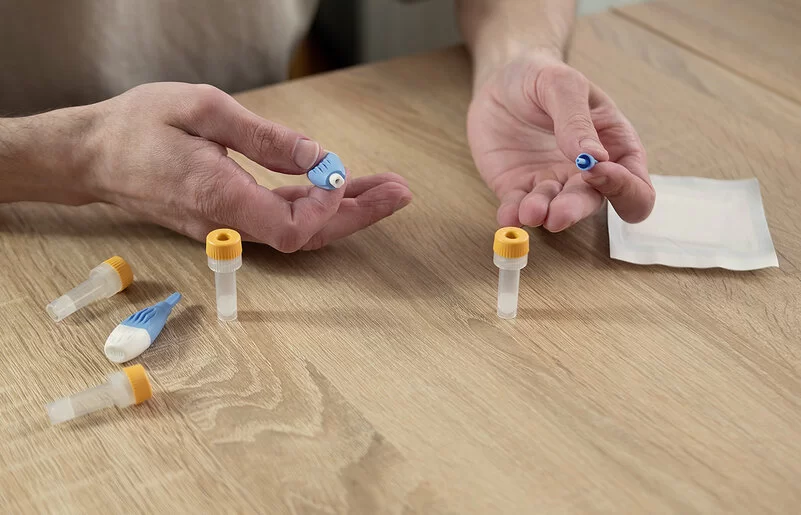 Die Hände eines Mannes liegen auf einer Tischplatte; auf dem Tisch liegen Utensilien für einen Blut-Selbsttest, darunter kleine Gefäße für Blutproben. Die Hände halten ein spezielles kleines Instrument zur Blutentnahme.