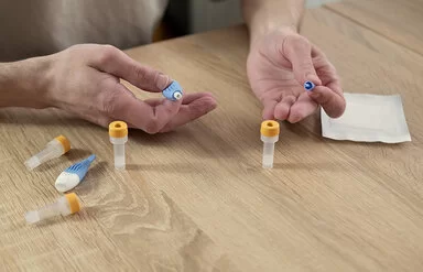 Die Hände eines Mannes liegen auf einer Tischplatte; auf dem Tisch liegen Utensilien für einen Blut-Selbsttest, darunter kleine Gefäße für Blutproben. Die Hände halten ein spezielles kleines Instrument zur Blutentnahme.
