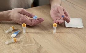 Die Hände eines Mannes liegen auf einer Tischplatte; auf dem Tisch liegen Utensilien für einen Blut-Selbsttest, darunter kleine Gefäße für Blutproben. Die Hände halten ein spezielles kleines Instrument zur Blutentnahme.