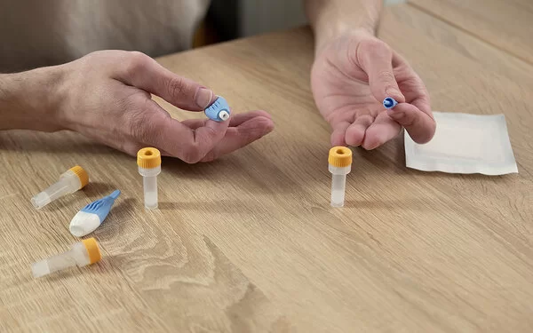 Die Hände eines Mannes liegen auf einer Tischplatte; auf dem Tisch liegen Utensilien für einen Blut-Selbsttest, darunter kleine Gefäße für Blutproben. Die Hände halten ein spezielles kleines Instrument zur Blutentnahme.