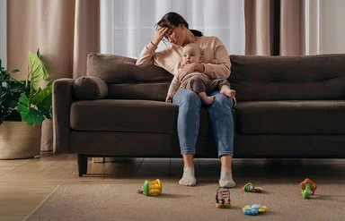 Eine junge Mutter sitzt mit ihrem Baby auf dem Schoss auf einer Couch in einem warm eingerichteten Wohnzimmer. Die Frau stützt ihr Kopf in die Hand, ihr Blick ist nach unten gerichtet. Auf dem Fußboden liegt vereinzelt Spielzeug.