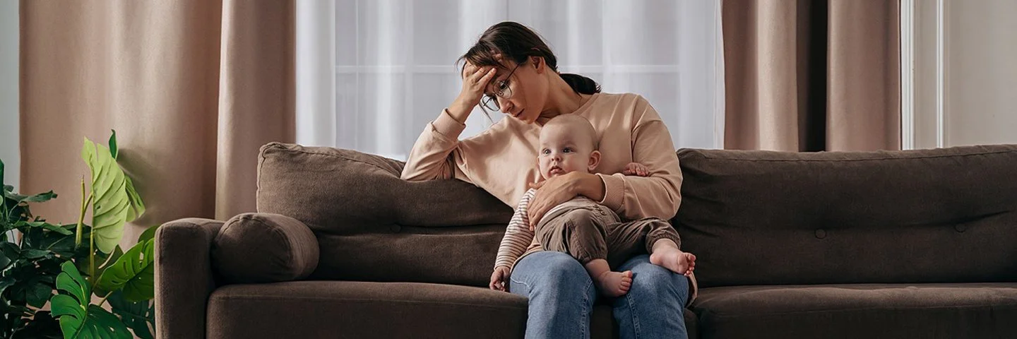 Eine junge Mutter sitzt mit ihrem Baby auf dem Schoss auf einer Couch in einem warm eingerichteten Wohnzimmer. Die Frau stützt ihr Kopf in die Hand, ihr Blick ist nach unten gerichtet. Auf dem Fußboden liegt vereinzelt Spielzeug.