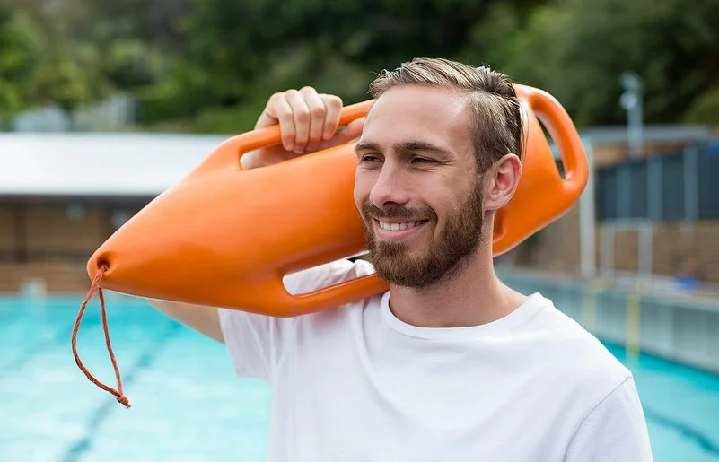 Ein lächelnder Mann in sportlichem T-Shirt mit einer Schwimmboje über der Schulter steht am Beckenrand eines Freibads.