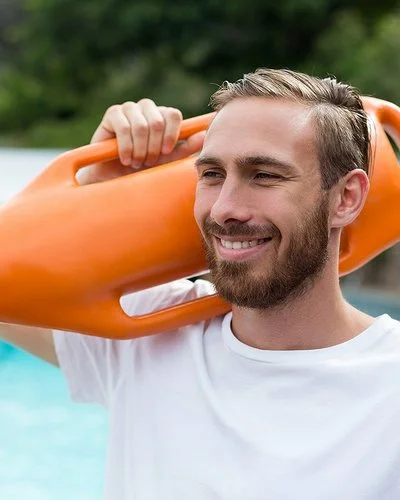 Ein lächelnder Mann in sportlichem T-Shirt mit einer Schwimmboje über der Schulter steht am Beckenrand eines Freibads.