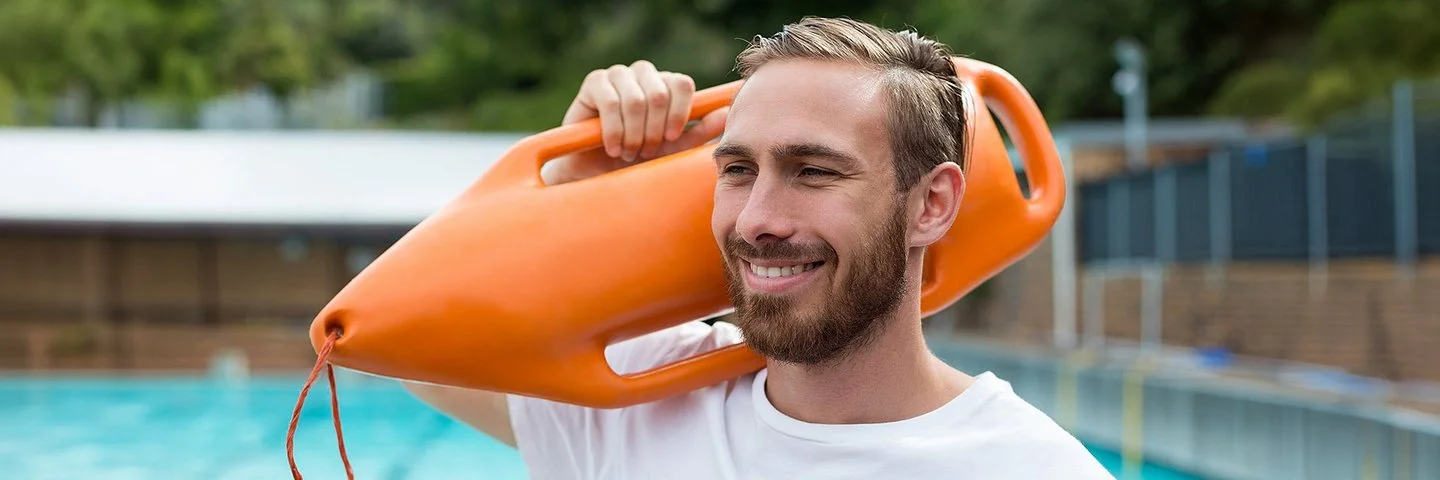 Ein lächelnder Mann in sportlichem T-Shirt mit einer Schwimmboje über der Schulter steht am Beckenrand eines Freibads.