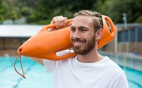 Ein lächelnder Mann in sportlichem T-Shirt mit einer Schwimmboje über der Schulter steht am Beckenrand eines Freibads.