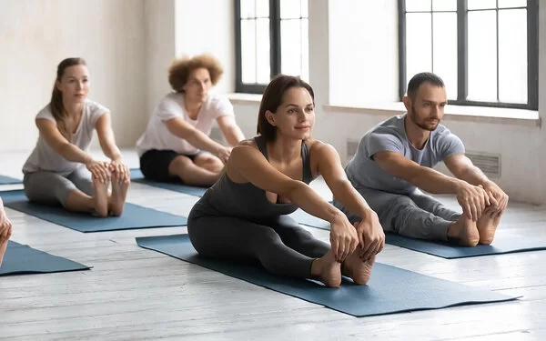 Mehrere Frauen und Männer machen zusammen einen Yin-Yoga-Kurs. Sie sitzen auf ihren Yogamatten und halten eine Dehnübung.