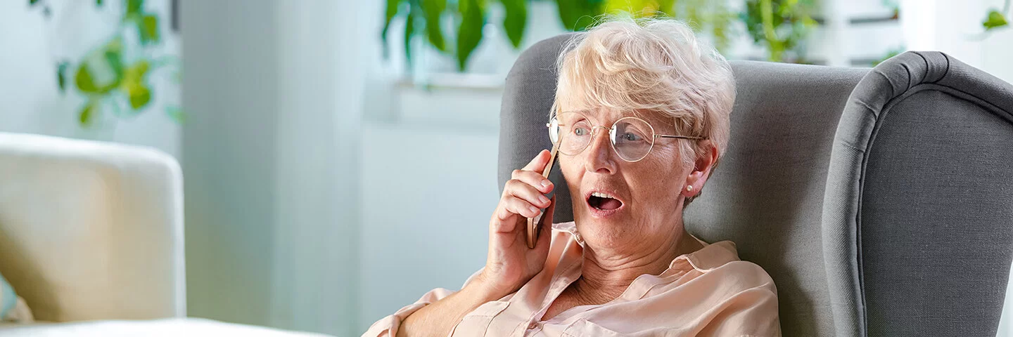 Ältere Frau sitzt mit einer Decke auf den Beinen in einem Sessel und telefoniert.