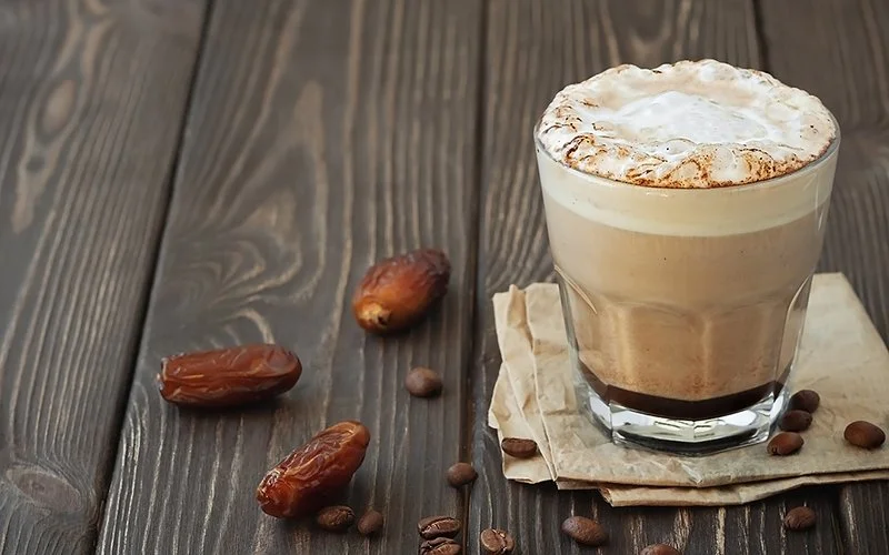 Ein Glas mit Cold Brew Kaffee steht auf einem Holztisch, daneben liegen Datteln und Kaffeebohnen.