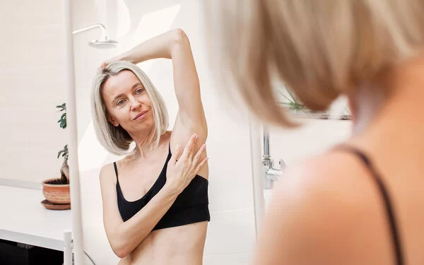 Eine Frau im Badezimmer vor dem Spiegel. Sie trägt eine schwarzes Bustier, hält einen Arm über den Kopf und tastet ihre Achselhöhle ab.
