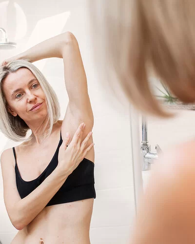Eine Frau im Badezimmer vor dem Spiegel. Sie trägt eine schwarzes Bustier, hält einen Arm über den Kopf und tastet ihre Achselhöhle ab.