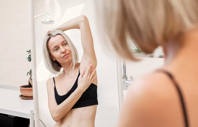 Eine Frau im Badezimmer vor dem Spiegel. Sie trägt eine schwarzes Bustier, hält einen Arm über den Kopf und tastet ihre Achselhöhle ab.