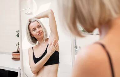 Eine Frau im Badezimmer vor dem Spiegel. Sie trägt eine schwarzes Bustier, hält einen Arm über den Kopf und tastet ihre Achselhöhle ab.