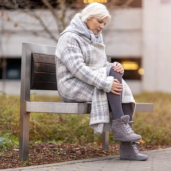 Eine Frau im mittleren Alter sitzt auf einer Bank in einer Grünanlage. Sie trägt einen dicken Wintermantel und reibt sich das Knie.
