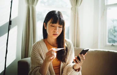 Eine junge Frau blickt auf ein Fieberthermometer in ihrer Hand. In der anderen Hand hält sie ein Smartphone.