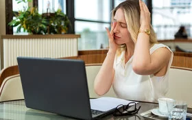Eine junge Frau sitzt vor ihrem Laptop und fasst sich mit schmerzverzerrtem Gesicht an ihr rechtes Auge und die linke Kopfseite.