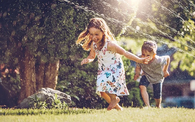Zwei Kinder laufen barfuß über eine Wiese und spielen lachend im sprühenden Wasser.