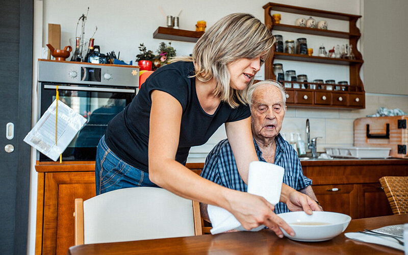 Eine junge Frau stellt einem älteren Mann mit Schluckstörungen einen Teller Suppe auf den Tisch.