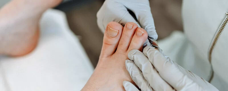 Eine Person trägt Handschuhe und schneidet die Fußnägel einer anderen Person, die entspannt auf einer Behandlungsbank liegt.
