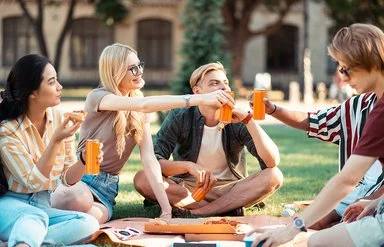 Eine Gruppe von fünf Jugendlichen in sommerlicher Kleidung sitzt im Kreis auf einer Wiese und stößt mit orangefarbenen Getränkedosen an.