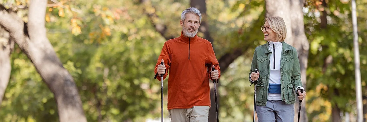 Ein Mann und eine Frau in sportlicher Freizeitkleidung beim Nordic-Walking in einem herbstlichen Park.