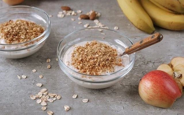 Auf grauem Hintergrund stehen zwei kleine Glasschalen, gefüllt mit Apfel-Zimt-Quark und Hafercrunch-Topping. Daneben sind zwei halbierte Äpfel arrangiert. Im Hintergrund verstreute Haferflocken und Mandeln sowie Bananen.