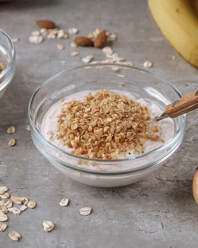 Auf grauem Hintergrund stehen zwei kleine Glasschalen, gefüllt mit Apfel-Zimt-Quark und Hafercrunch-Topping. Daneben sind zwei halbierte Äpfel arrangiert. Im Hintergrund verstreute Haferflocken und Mandeln sowie Bananen.