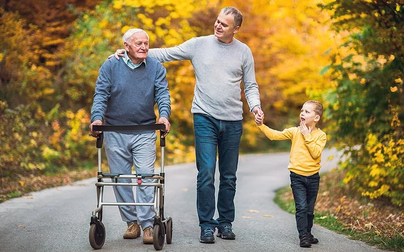 Ein alter Mann mit Rollator, ein Mann mittleren Alters und ein Junge gehen nebeneinander auf einem Weg durch einen herbstlichen Wald.
