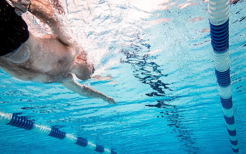 Unterwasseraufnahme eines Mannes mit einem Stomabeutel, der in einem Schwimmbecken krault.