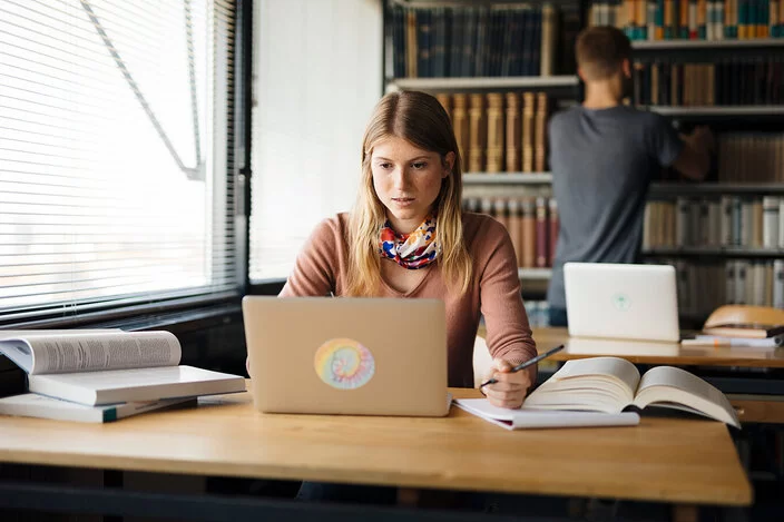 Eine junge, blonde Frau sitzt an einem Schreibtisch und tippt etwas in einen Laptop. Hinter ihr steht ein Mann und räumt etwas in ein Regal hinein.