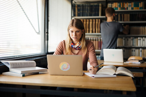 Eine junge, blonde Frau sitzt an einem Schreibtisch und tippt etwas in einen Laptop. Hinter ihr steht ein Mann und räumt etwas in ein Regal hinein.