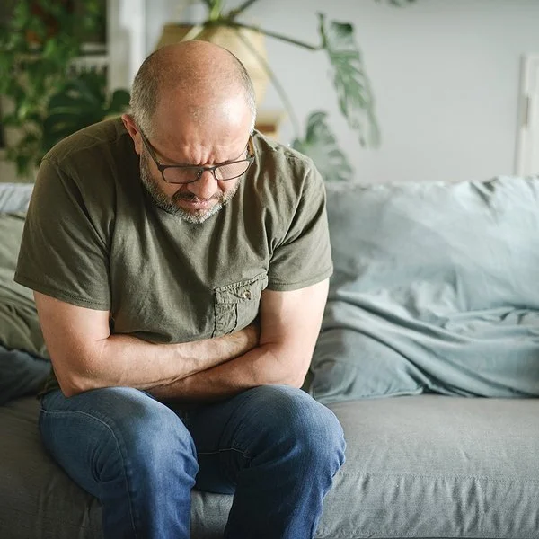 Ein Mann mit schmerzverzerrtem Gesicht sitzt auf einer Couch und hält sich mit beiden Händen den Magen.