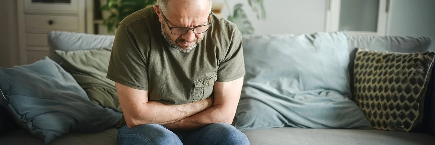 Ein Mann mit schmerzverzerrtem Gesicht sitzt auf einer Couch und hält sich mit beiden Händen den Magen.
