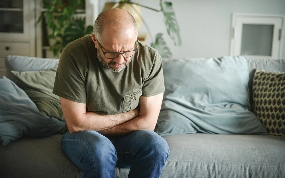Ein Mann mit schmerzverzerrtem Gesicht sitzt auf einer Couch und hält sich mit beiden Händen den Magen.