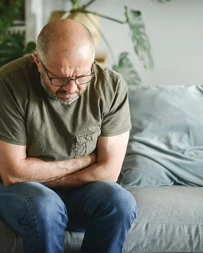 Ein Mann mit schmerzverzerrtem Gesicht sitzt auf einer Couch und hält sich mit beiden Händen den Magen.