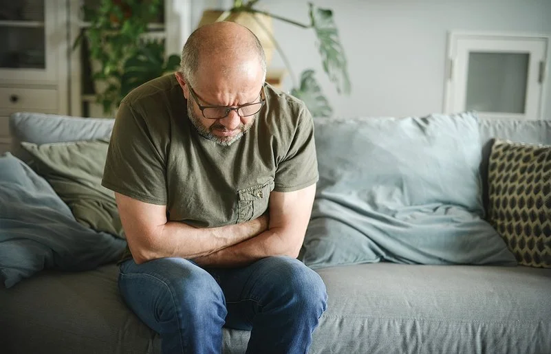 Ein Mann mit schmerzverzerrtem Gesicht sitzt auf einer Couch und hält sich mit beiden Händen den Magen.