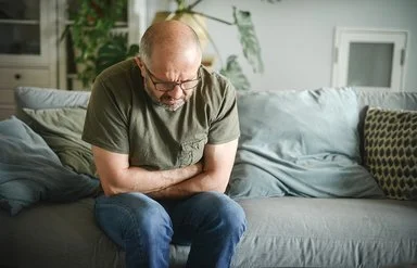 Ein Mann mit schmerzverzerrtem Gesicht sitzt auf einer Couch und hält sich mit beiden Händen den Magen.