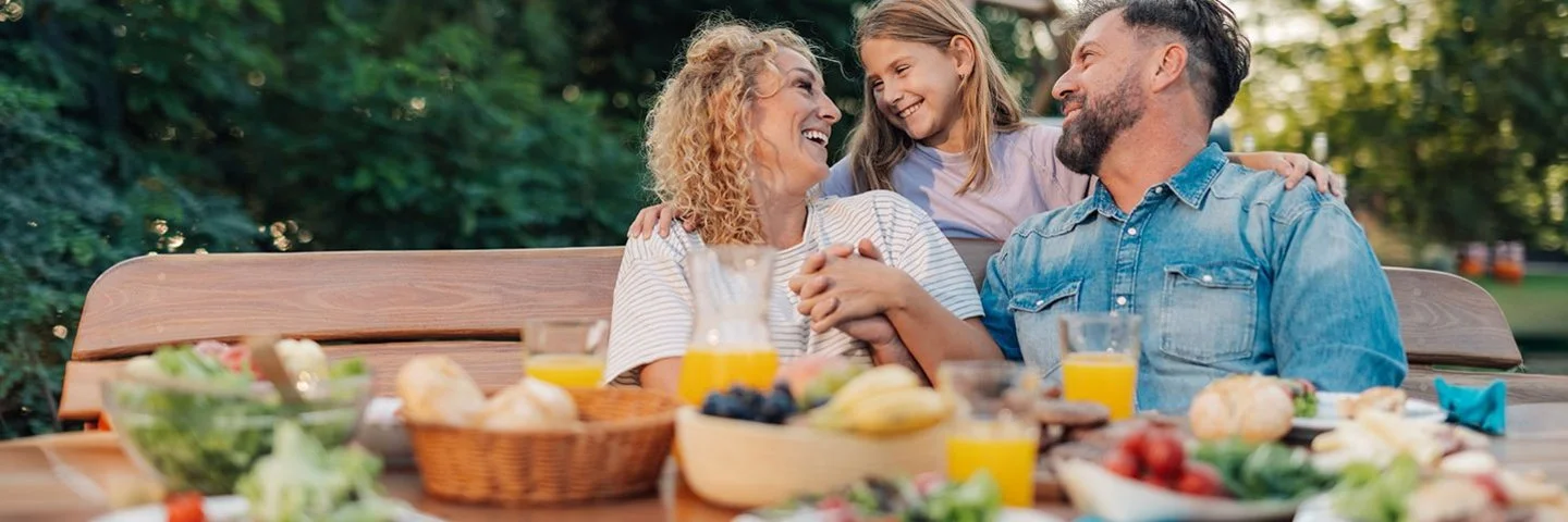 Eine Familie sitzt lachend an einem Gartentisch mit verschiedenen Speisen und Getränken im Freien