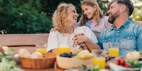 Eine Familie sitzt lachend an einem Gartentisch mit verschiedenen Speisen und Getränken im Freien