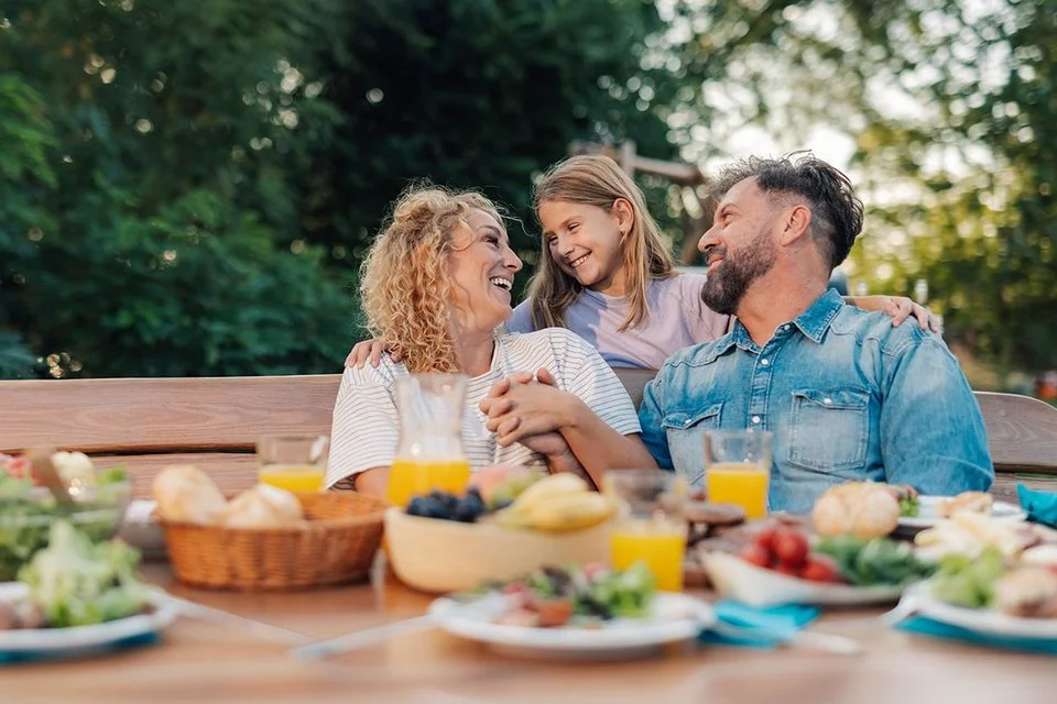 Eine Familie sitzt lachend an einem Gartentisch mit verschiedenen Speisen und Getränken im Freien