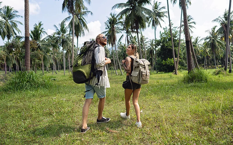 Zwei Backpacker laufen mit großen Rucksäcken und kurzen Hosen durch einen Palmenhain.