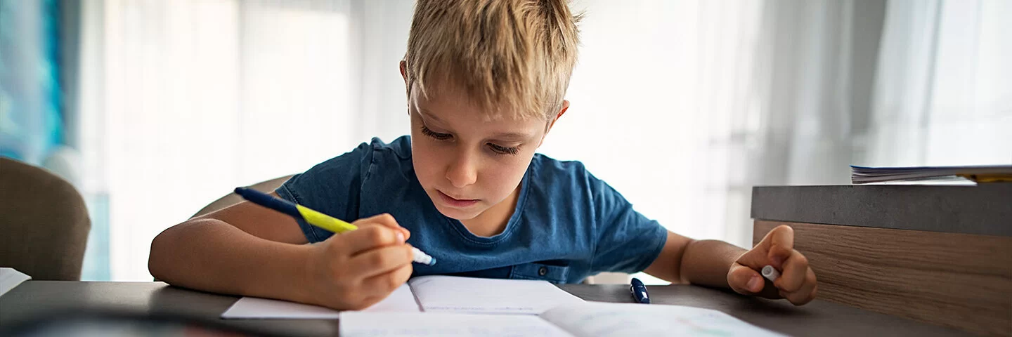 Ein etwa siebenjähriger Junge sitzt an seinem Schreibtisch und macht Hausaufgaben.