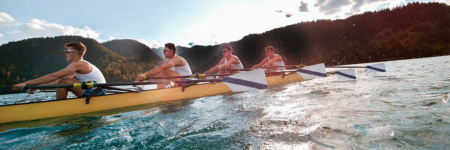 Vier Männer in sportlicher Kleidung rudern in einem Ruderboot auf einem See.