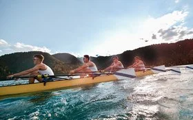 Vier Männer in sportlicher Kleidung rudern in einem Ruderboot auf einem See.