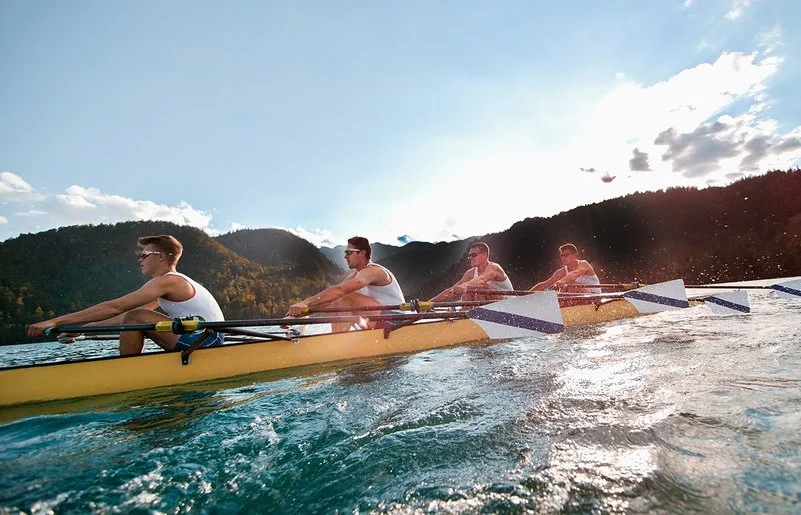 Vier Männer in sportlicher Kleidung rudern in einem Ruderboot auf einem See.
